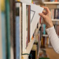 Female hand pulling book from bookshelf in public library in university, college or high school. Woman student take novel from bookcase in bookshop store, soft focus. Education and literature concept